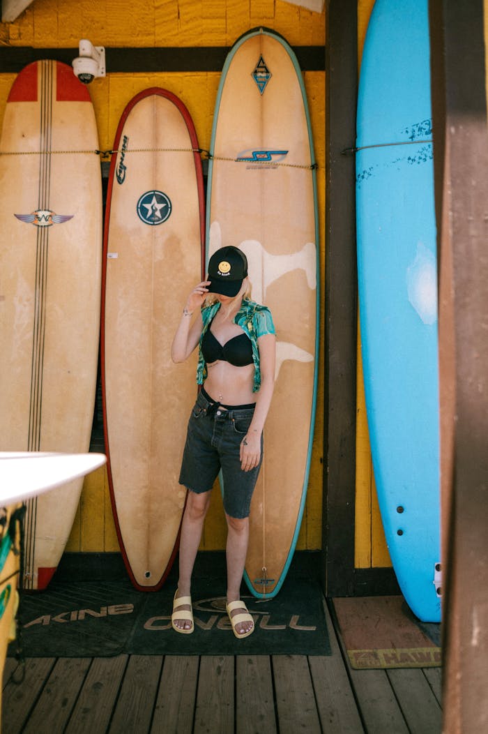 A young woman in summer attire posing with surfboards in an outdoor surf shop setting.